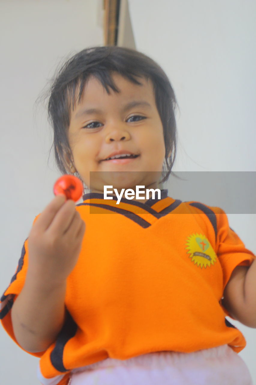 PORTRAIT OF CUTE BOY HOLDING ORANGE WHILE STANDING AGAINST WALL