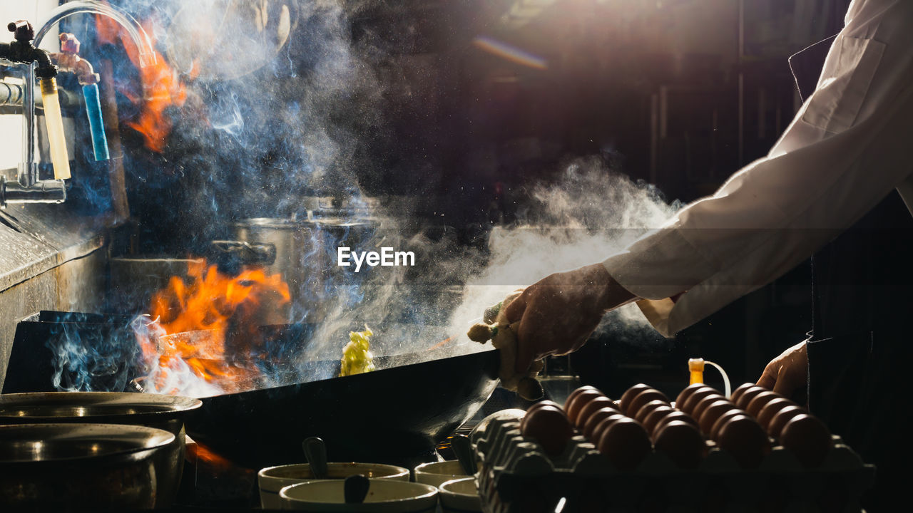 Chef stir fry the food in a frying pan, smoke and splatter the sauce in the kitchen.