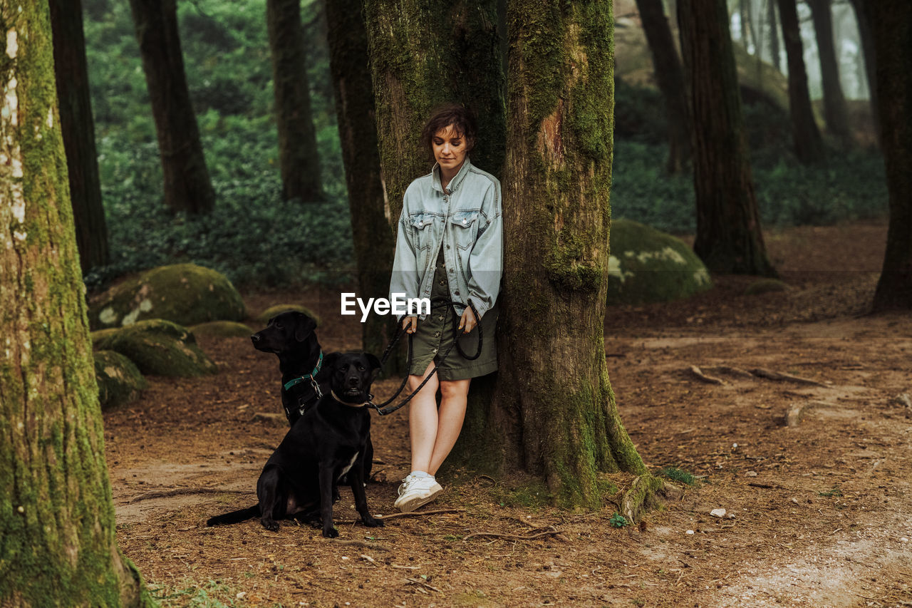 Woman with two dogs in the forest leaning against a mossy tree