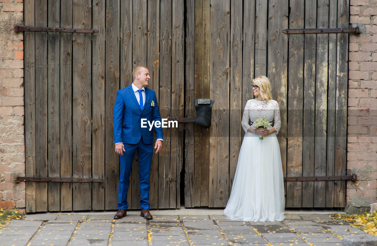 Smiling bridal couple looking at each other while standing against door of barn