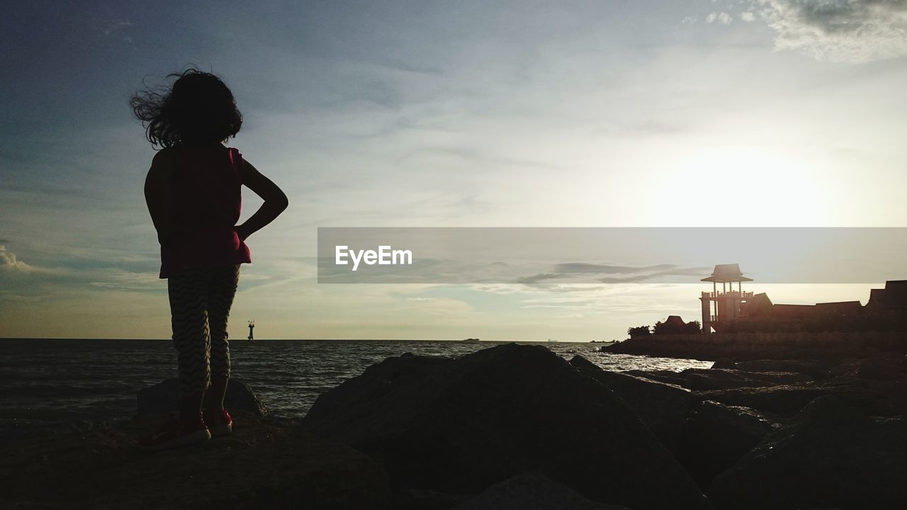 FULL LENGTH OF BOY STANDING ON ROCK AT SHORE AGAINST SKY