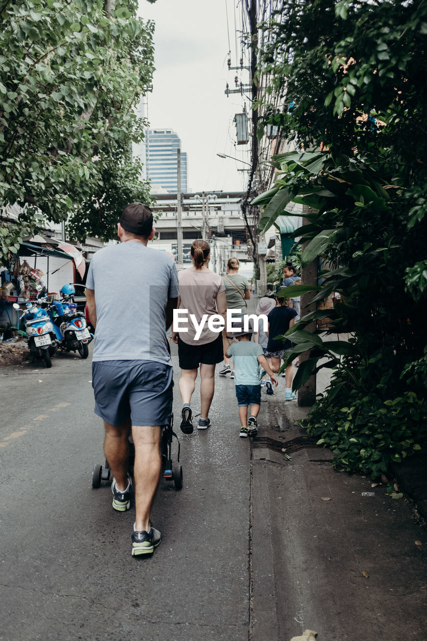 REAR VIEW OF PEOPLE WALKING ON ROAD ALONG TREES