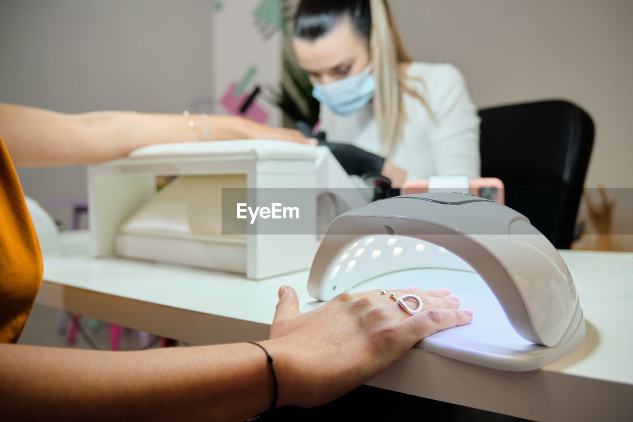 Crop anonymous female customer putting hand on uv lamp for drying nails while getting professional manicure in modern salon