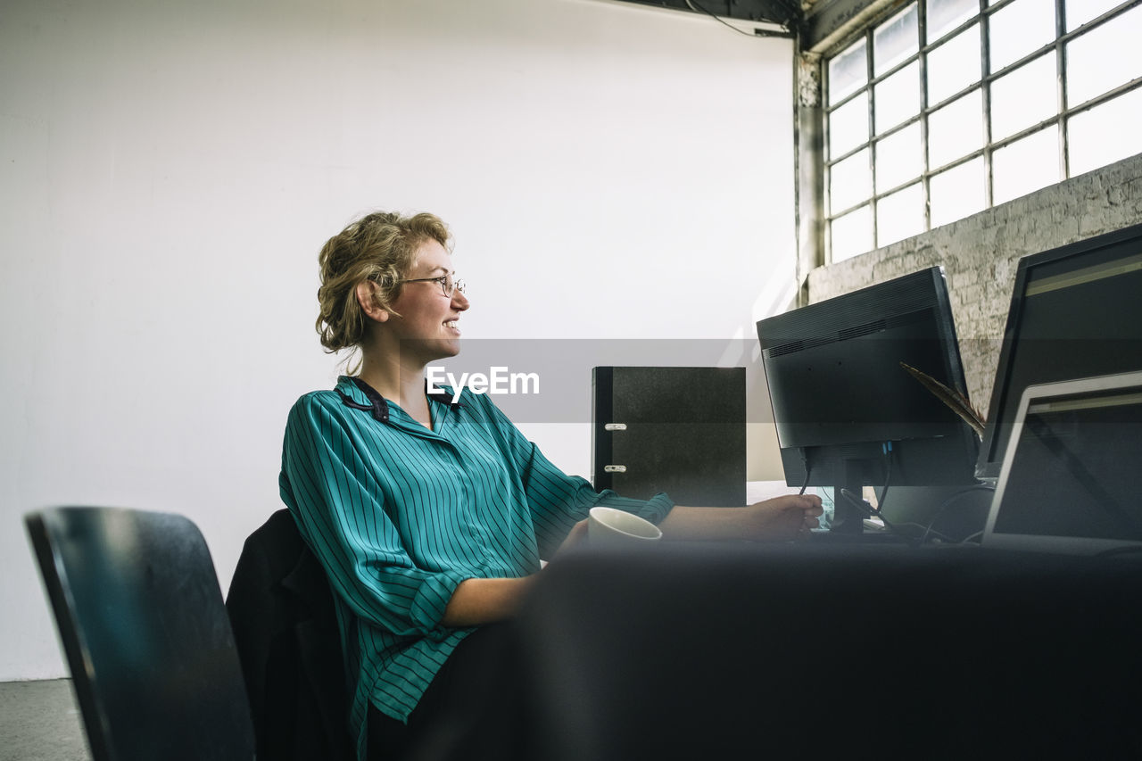 Thoughtful young businesswoman smiling and looking away while sitting at desk in creative office