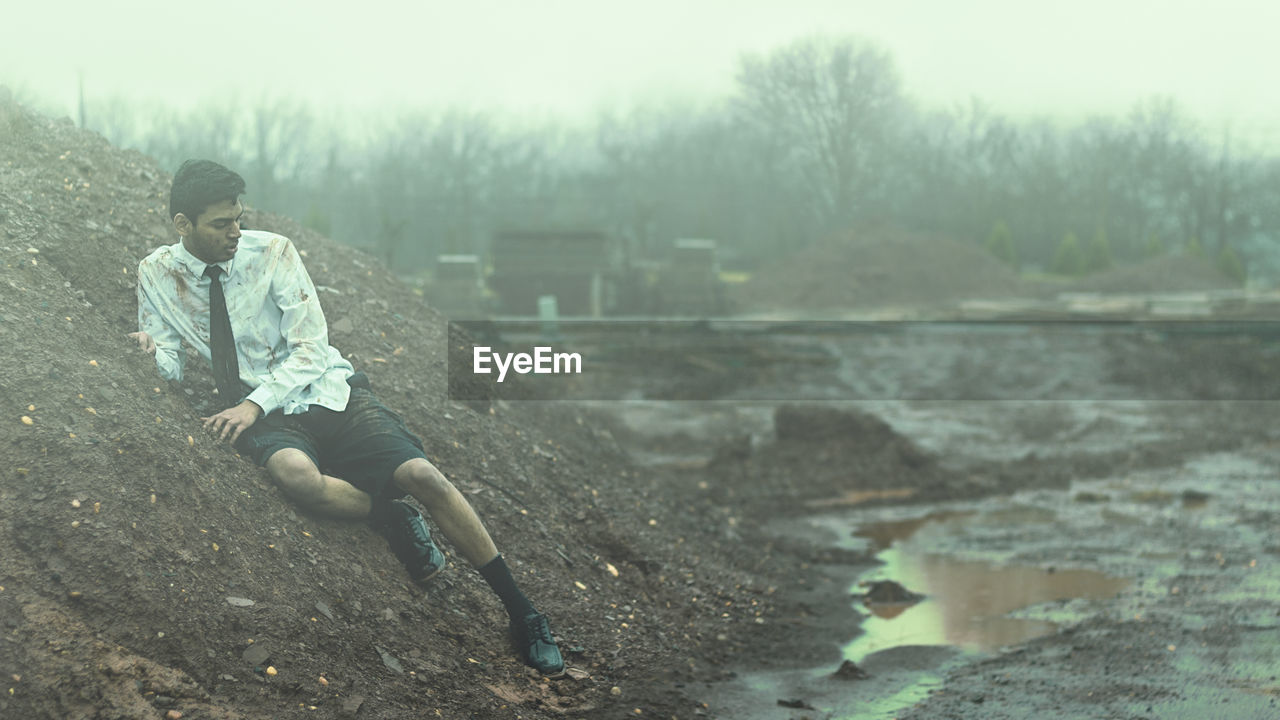 Young man sitting on mud against sky