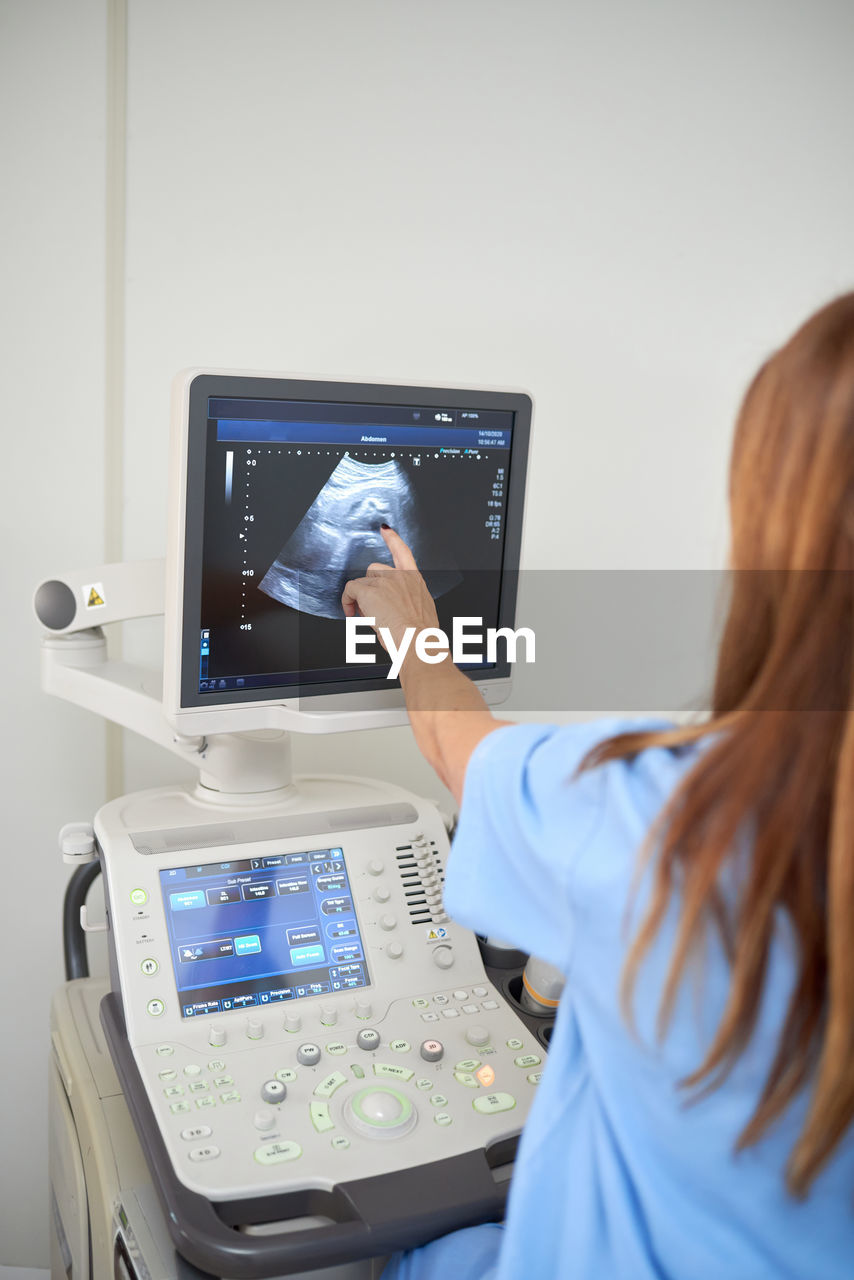 Back view of crop unrecognizable doctor showing video on ultrasound machine display during sonography diagnostic to patient