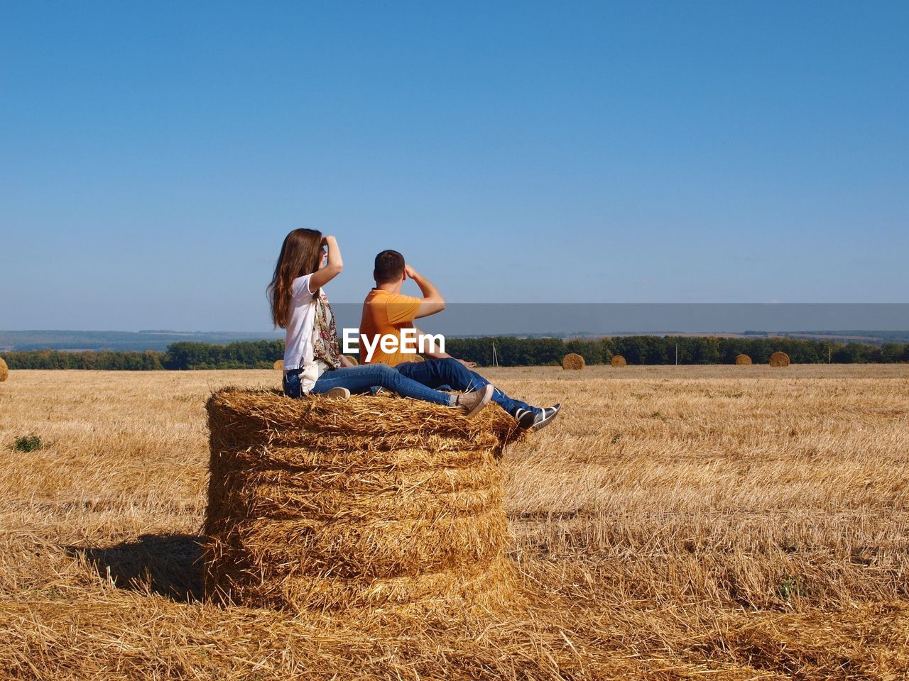 Side view of couple sitting on hay bale against clear sky