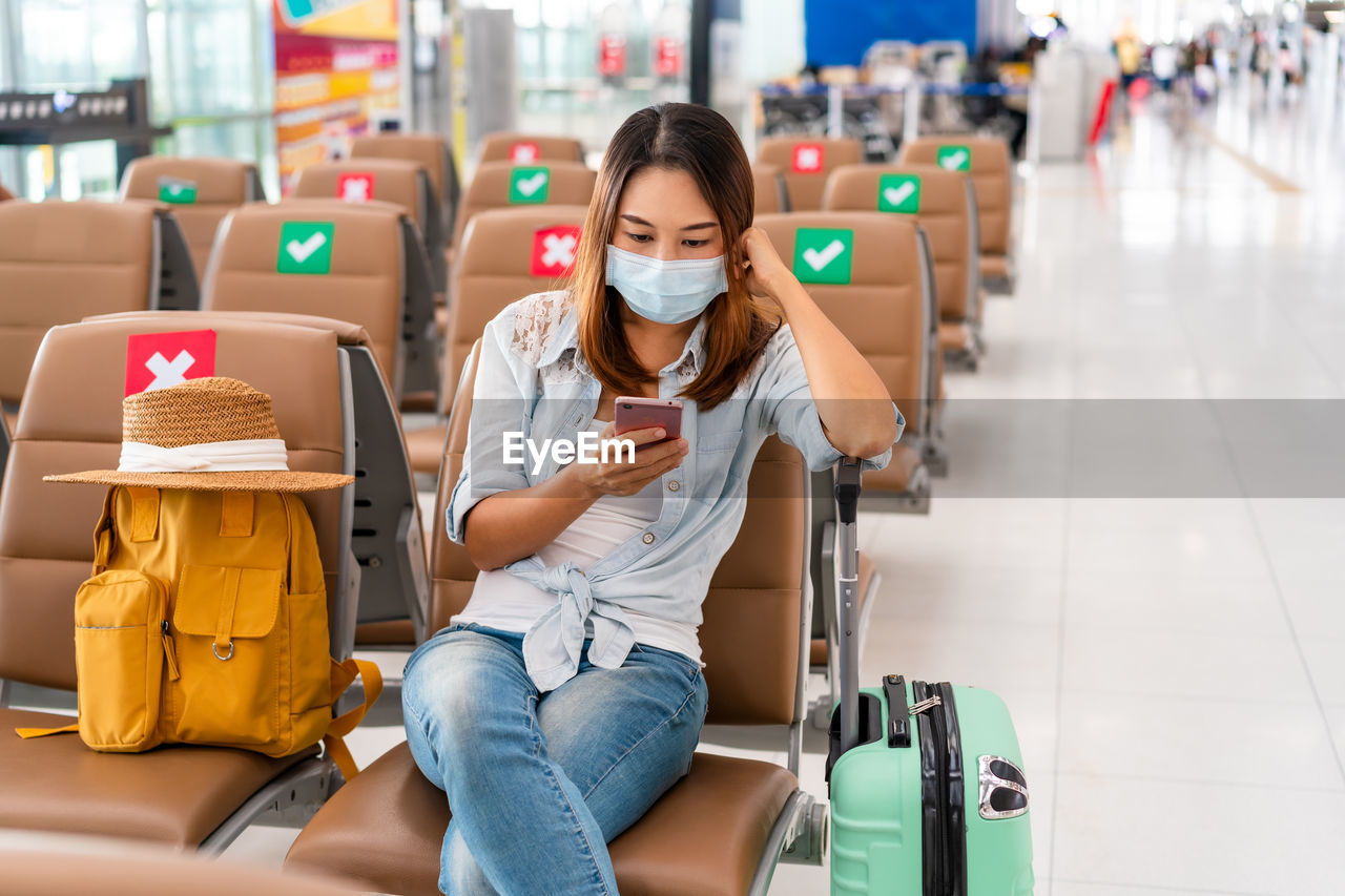 Woman wearing mask using mobile phone while sitting at airport