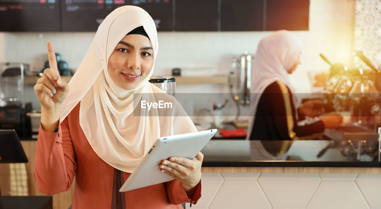 PORTRAIT OF SMILING YOUNG WOMAN HOLDING CAMERA WHILE STANDING AGAINST INDOORS