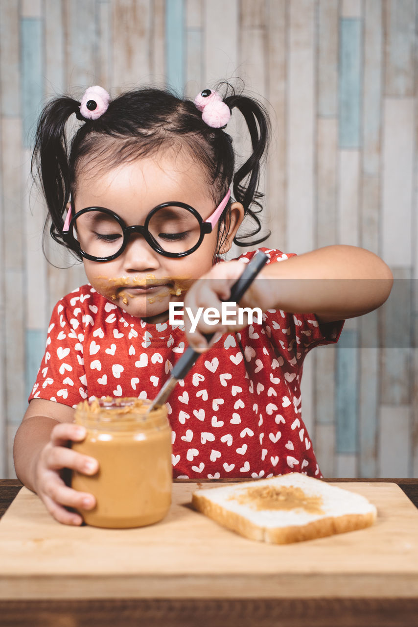 Girl applying peanut butter on bread at table against wall