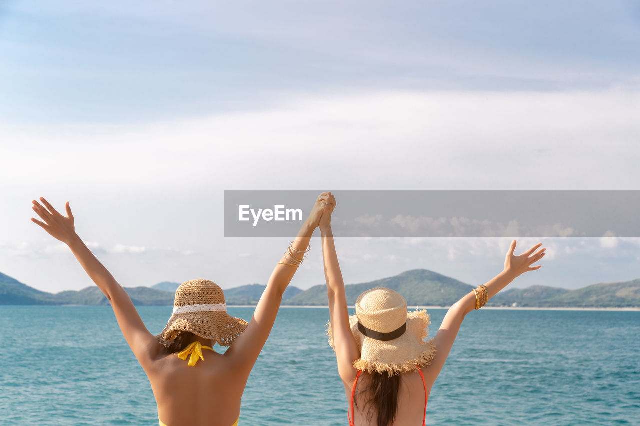 Rear view of young friends wearing hat with arms raised on beach