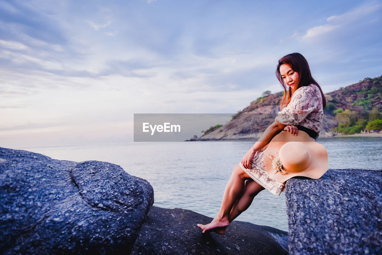 WOMAN SITTING ON ROCK AT SEA AGAINST SKY