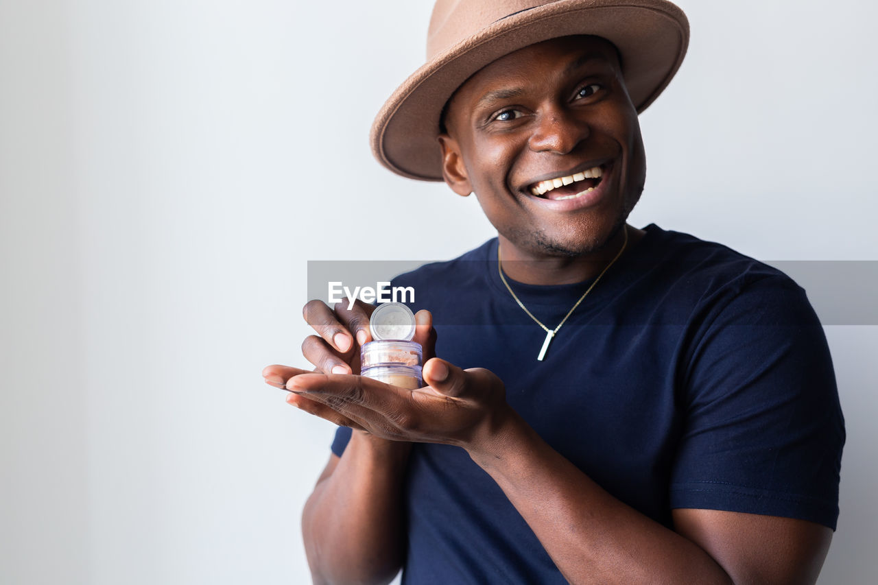 one person, smiling, happiness, portrait, adult, hat, person, emotion, clothing, men, cheerful, studio shot, looking at camera, indoors, casual clothing, fashion accessory, holding, lifestyles, young adult, cowboy hat, waist up, smile, teeth, positive emotion, copy space, enjoyment, sun hat, front view, standing, relaxation, looking, t-shirt, human face