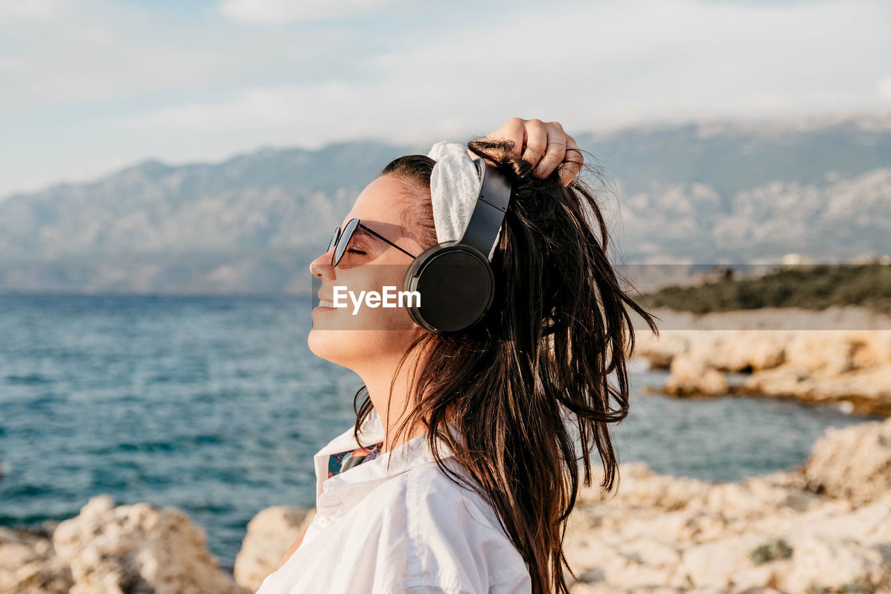 Side view of young woman in white shirt listening to music on headphones. summer, beach, lifestyle.