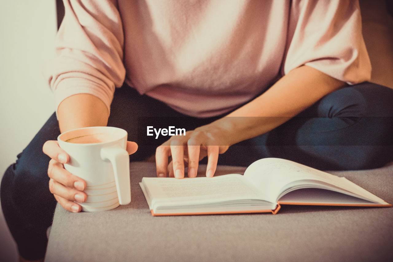 Unrecognizable woman drinking coffee and reading book while relaxing at home.