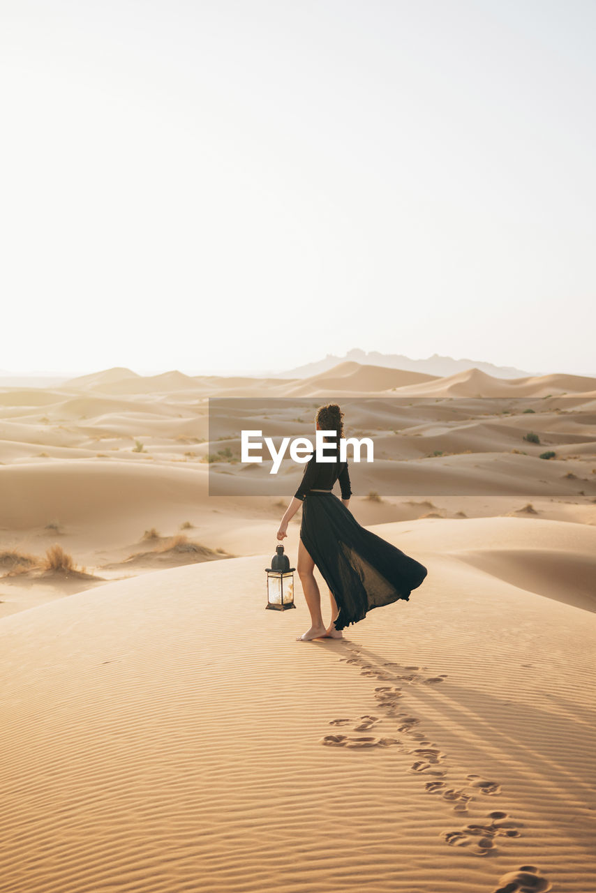 Side view of woman holding lantern while standing at sahara desert against clear sky during sunset