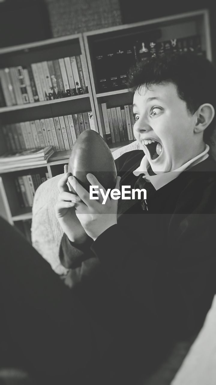 Boy with mouth open looking at easter egg while sitting against shelf