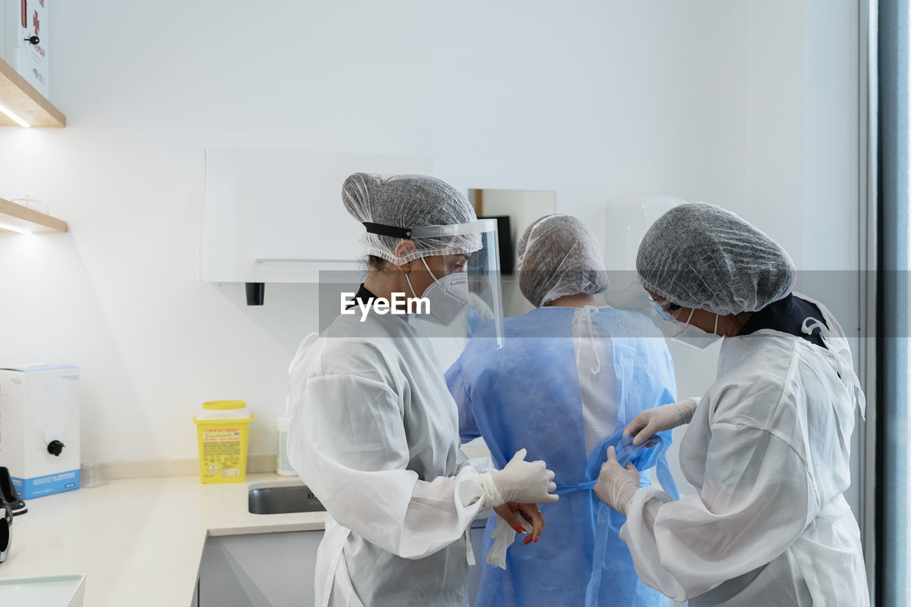 Female medical workers in protective masks and face shields helping each other to get dressed while preparing for medical procedure in hospital