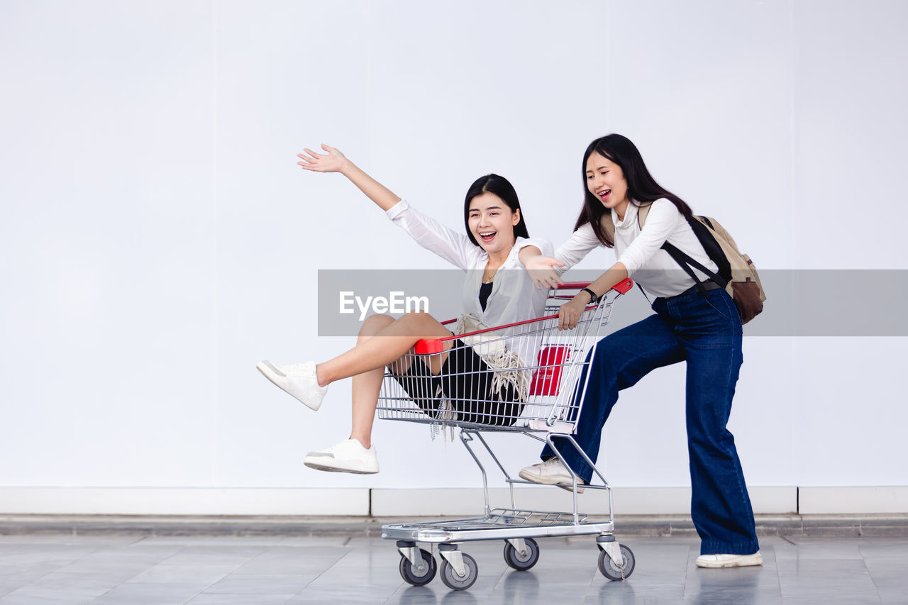 Smiling young woman sitting in shopping cart with friend pushing on tiled floor