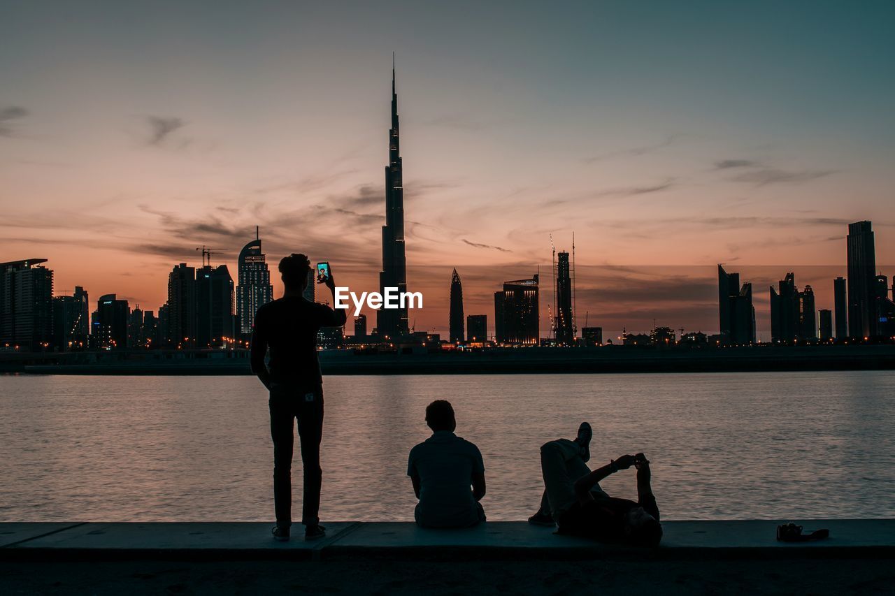 People on retaining wall by river with cityscape in background during sunset