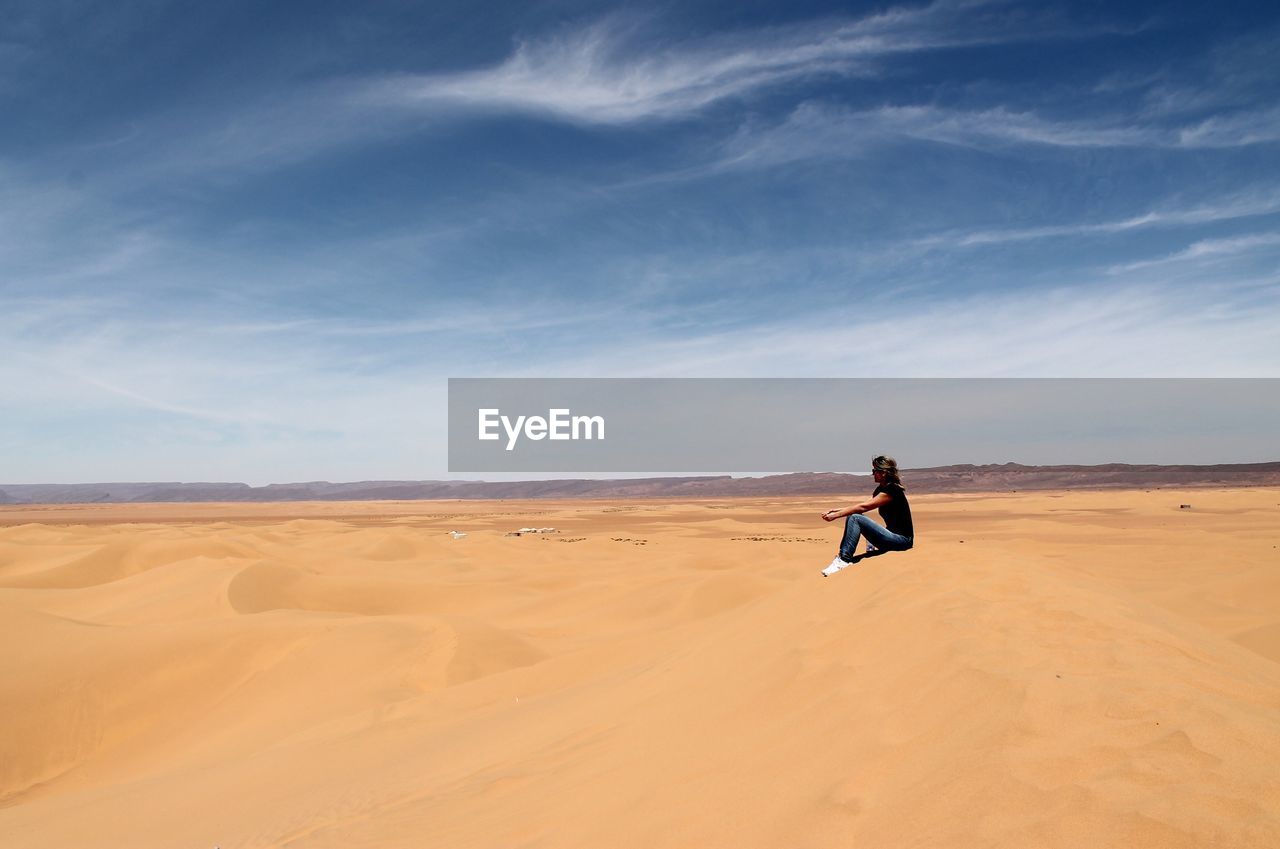 Full length of woman sitting on sand dune at sahara desert against sky