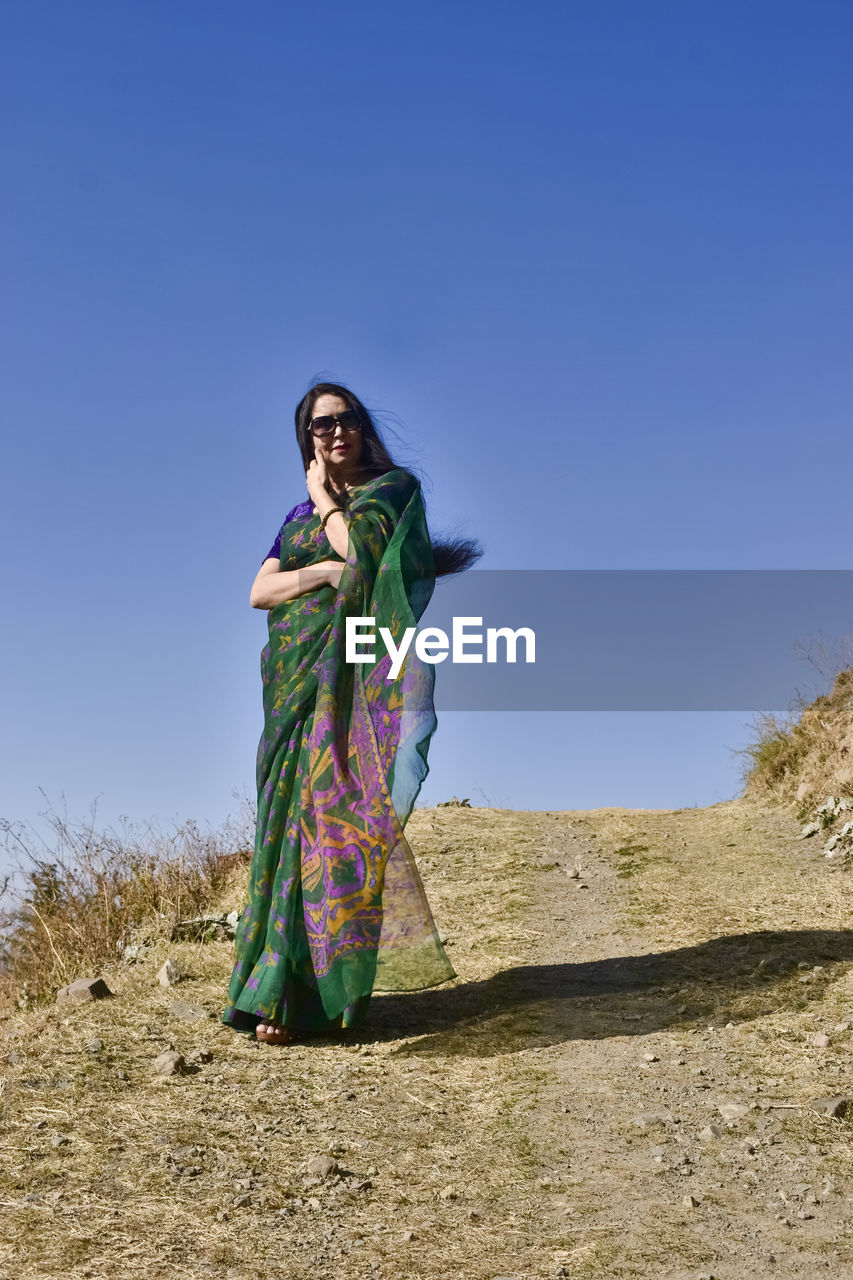 one person, adult, traditional clothing, clothing, sky, nature, full length, blue, women, clear sky, land, standing, sunny, young adult, copy space, sunlight, landscape, fashion, day, outdoors, tradition, front view, rural scene, portrait, looking, person, female, environment, lifestyles, desert, smiling, sand, plant, holding, leisure activity, travel, looking away, dress