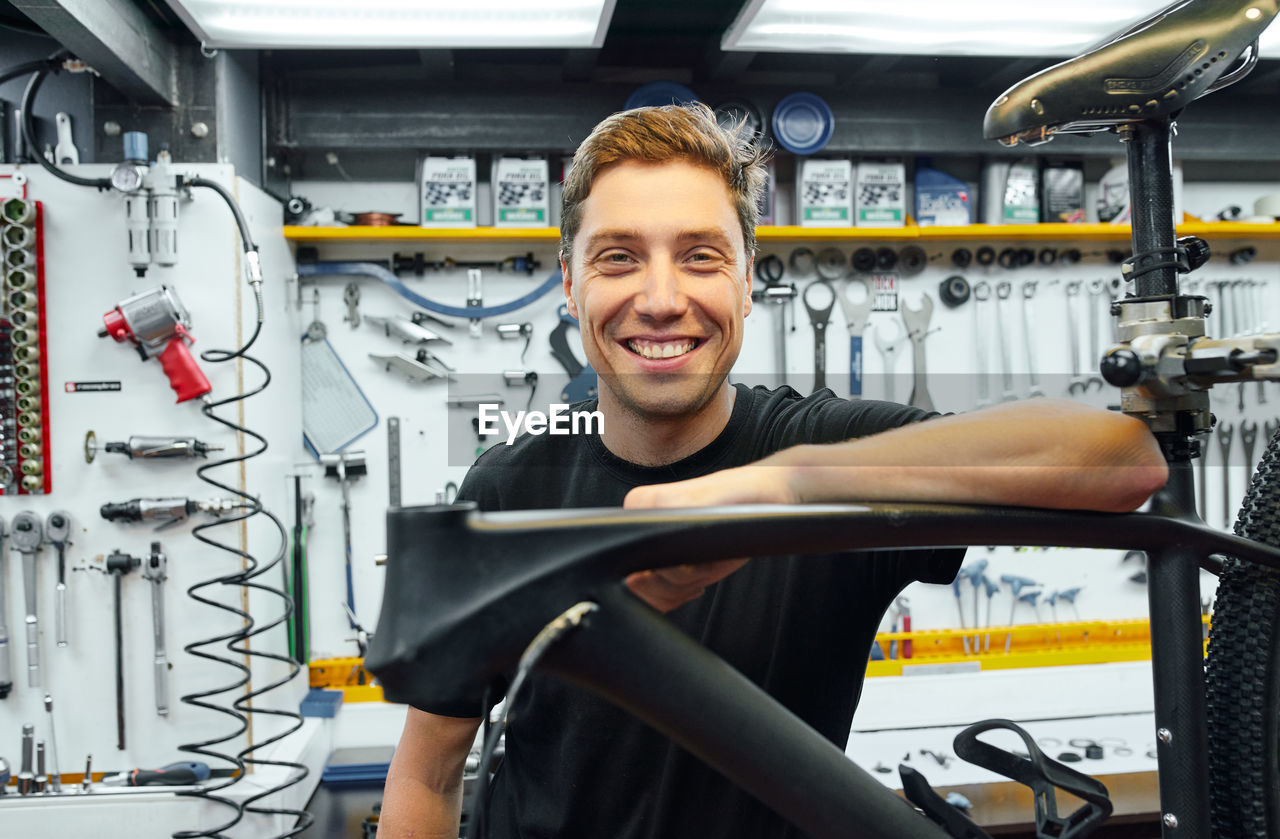 Happy male mechanic smiling and looking at camera while leaning on bike under repair against wall with tools in garage