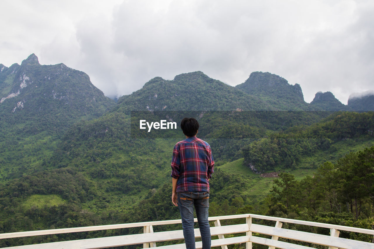 Rear view of man standing by railing against mountains and sky