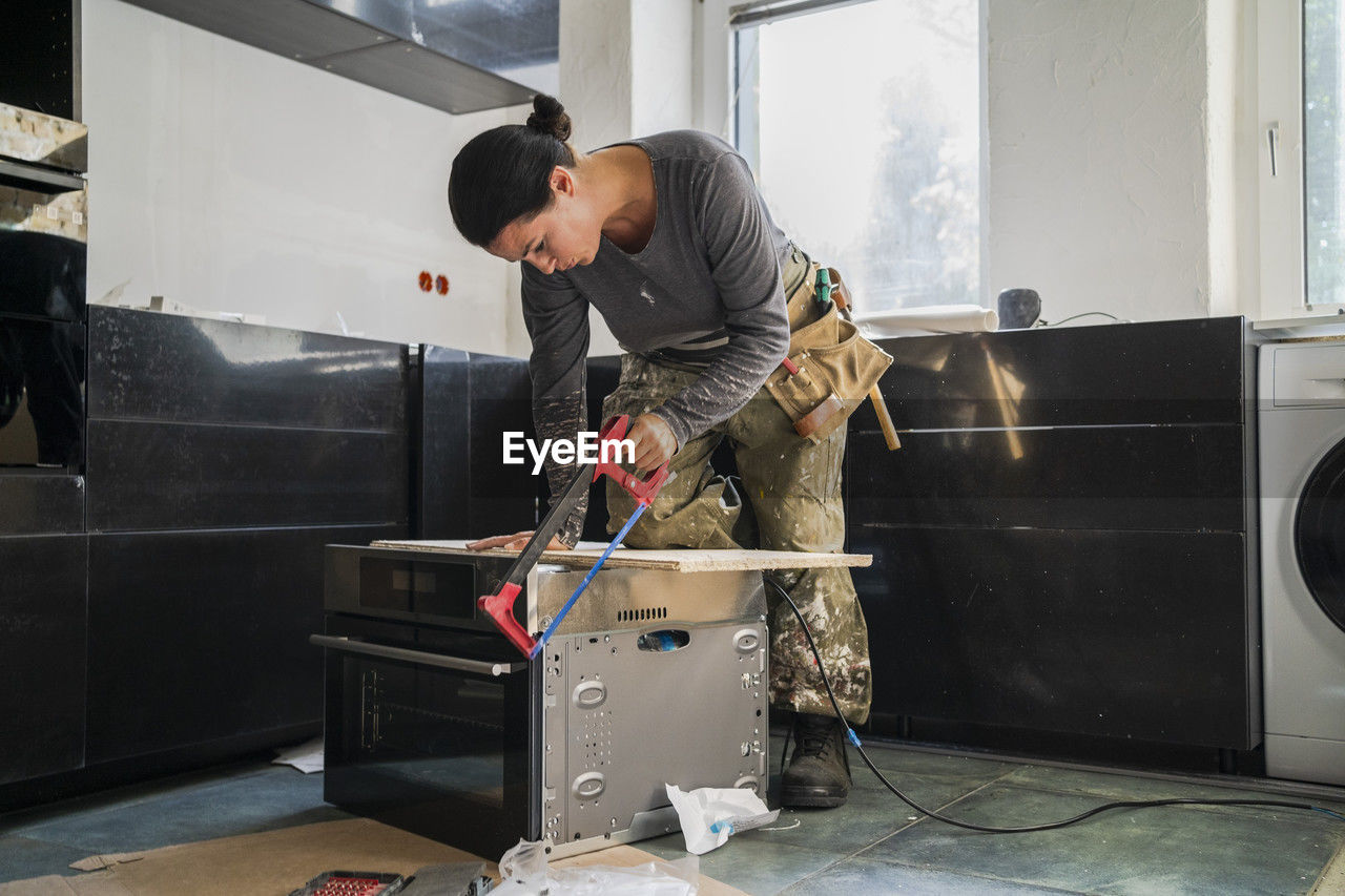 Female carpenter cutting plank on microwave in kitchen at apartment