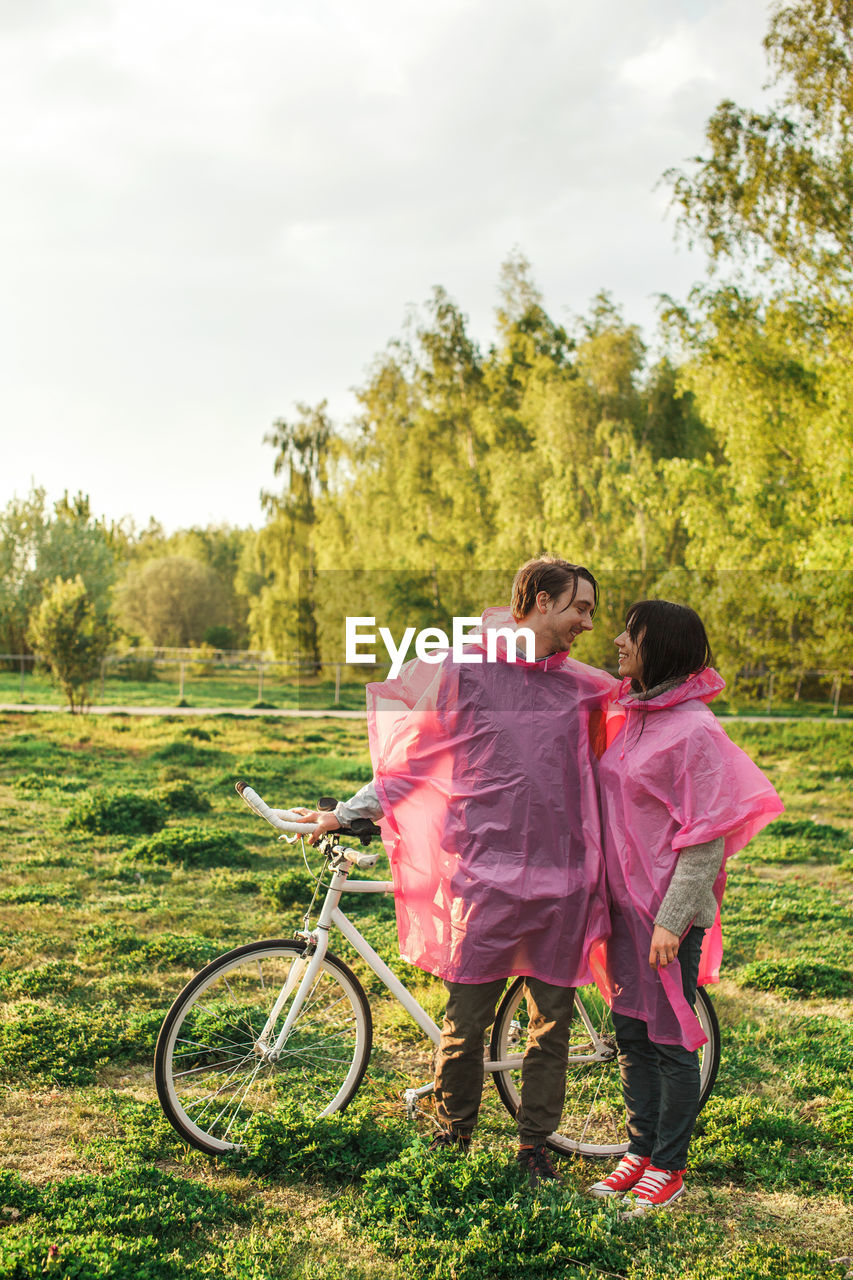 Young couple in pink raincoat standing with bicycle on field against sky