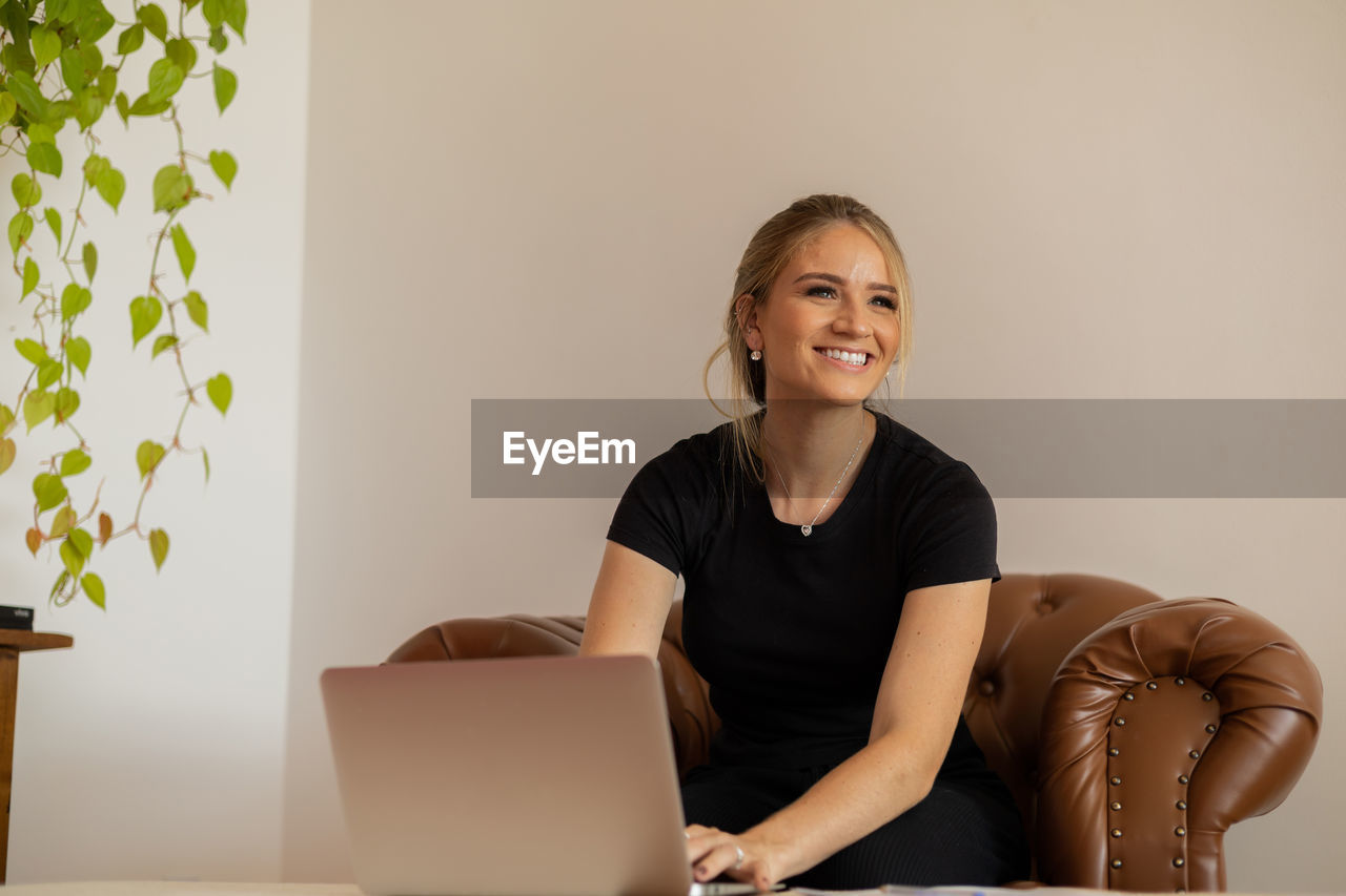 adult, one person, laptop, smiling, indoors, using laptop, technology, computer, wireless technology, women, happiness, sitting, communication, portrait, business, person, furniture, working, emotion, looking at camera, female, lifestyles, business finance and industry, businesswoman, cheerful, internet, copy space, table, computer network, front view, home interior, occupation, office, clothing, casual clothing, hairstyle, young adult, conversation, success, living room, relaxation, convenience, looking, brown hair, enjoyment