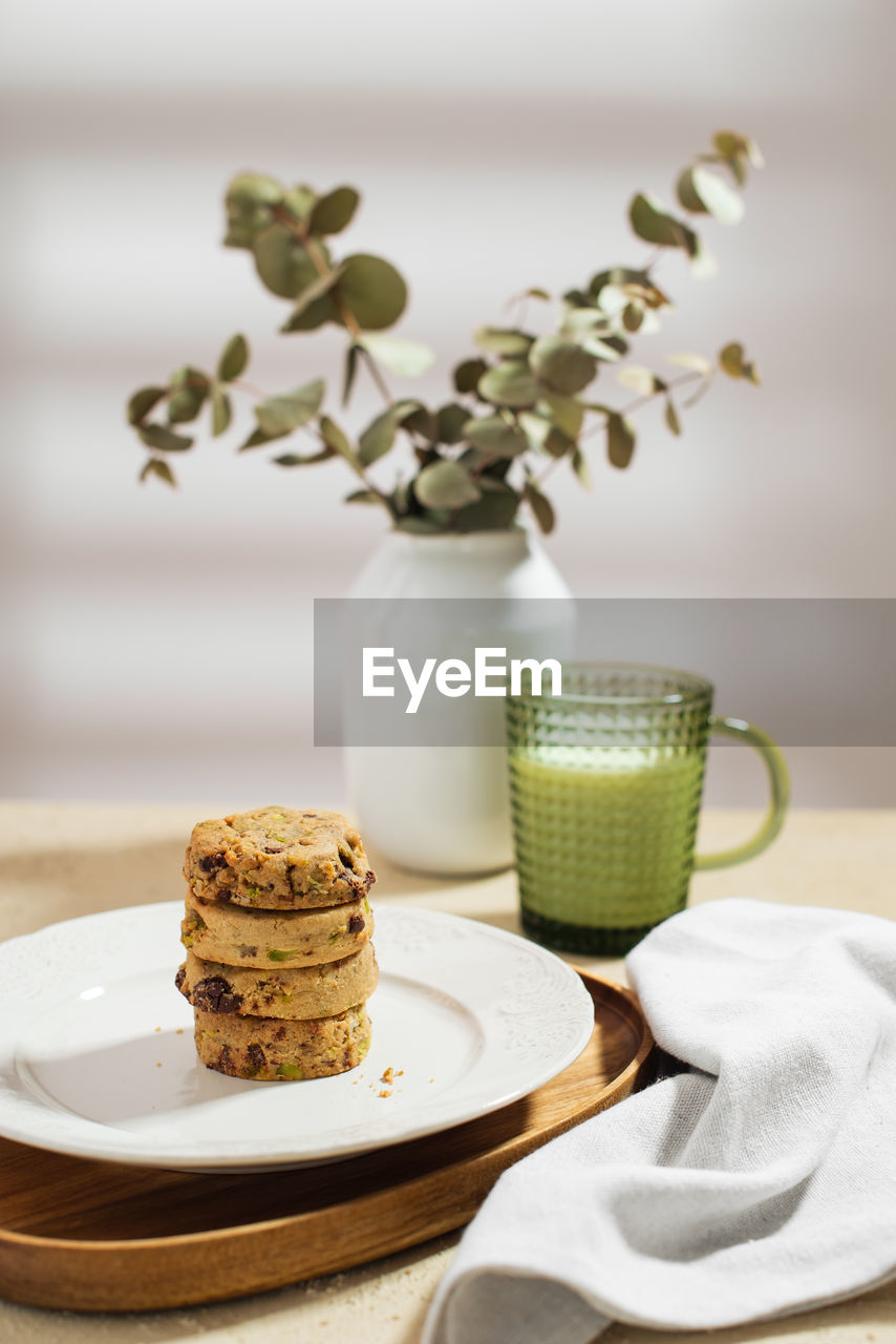 Plate with stack of tasty biscuits and mug of fresh milk placed on table near napkin and vase with twigs in morning