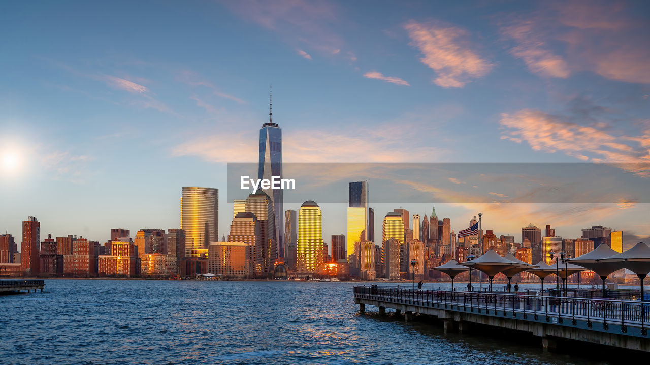 modern buildings by river against sky at sunset