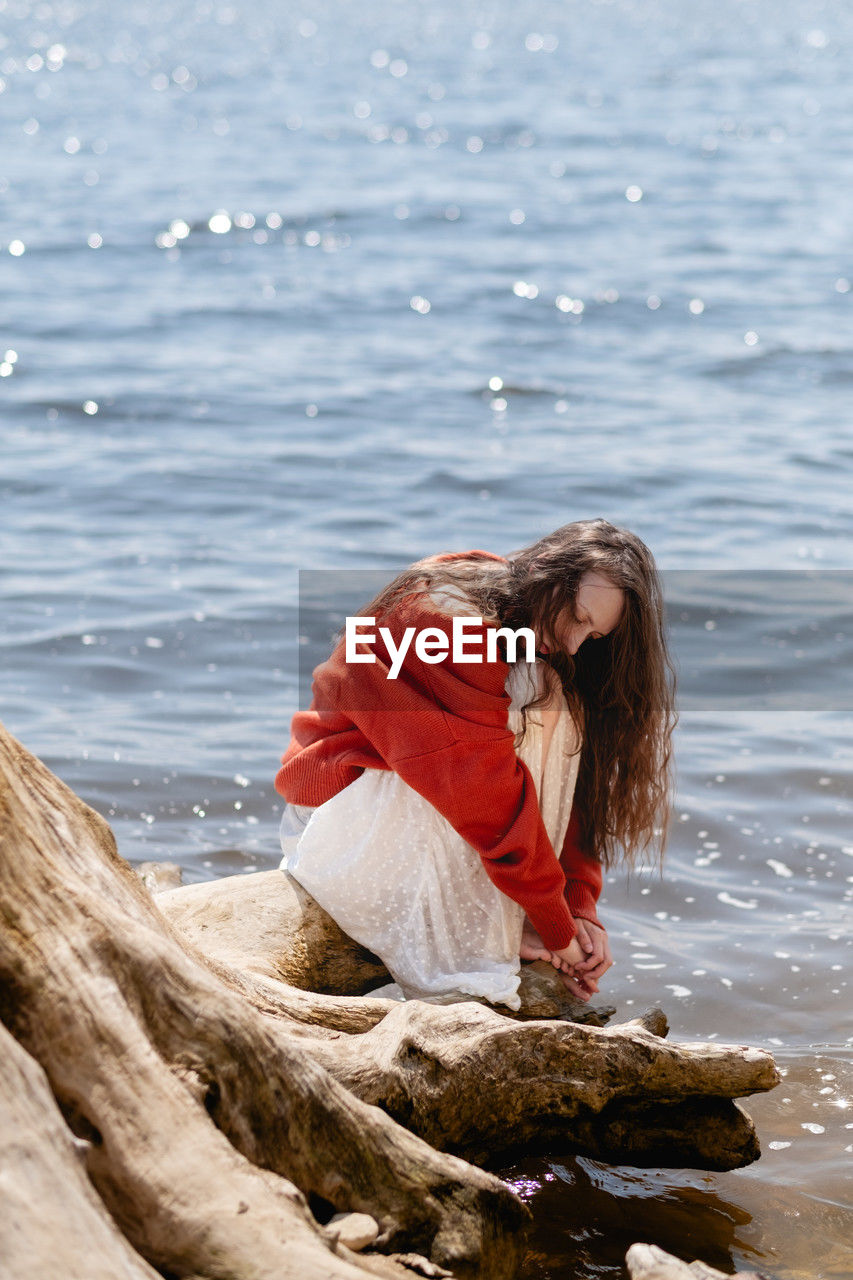 Woman with long hair and closed eyes, in a terracotta sweater, sits on a tree stump