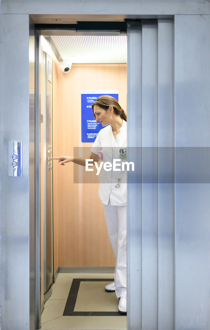 Full body woman in white medical uniform pushing button inside elevator while working in hospital