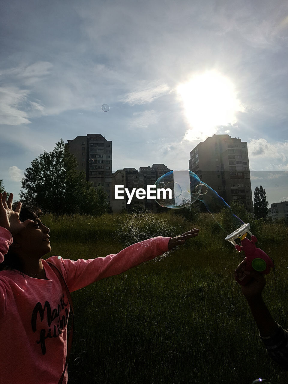 Sisters playing with bubble gun on grassy field by buildings against sky