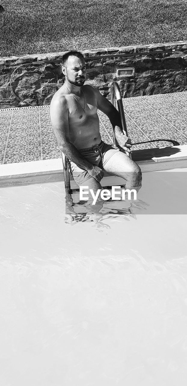 PORTRAIT OF YOUNG MAN SITTING ON SWIMMING POOL