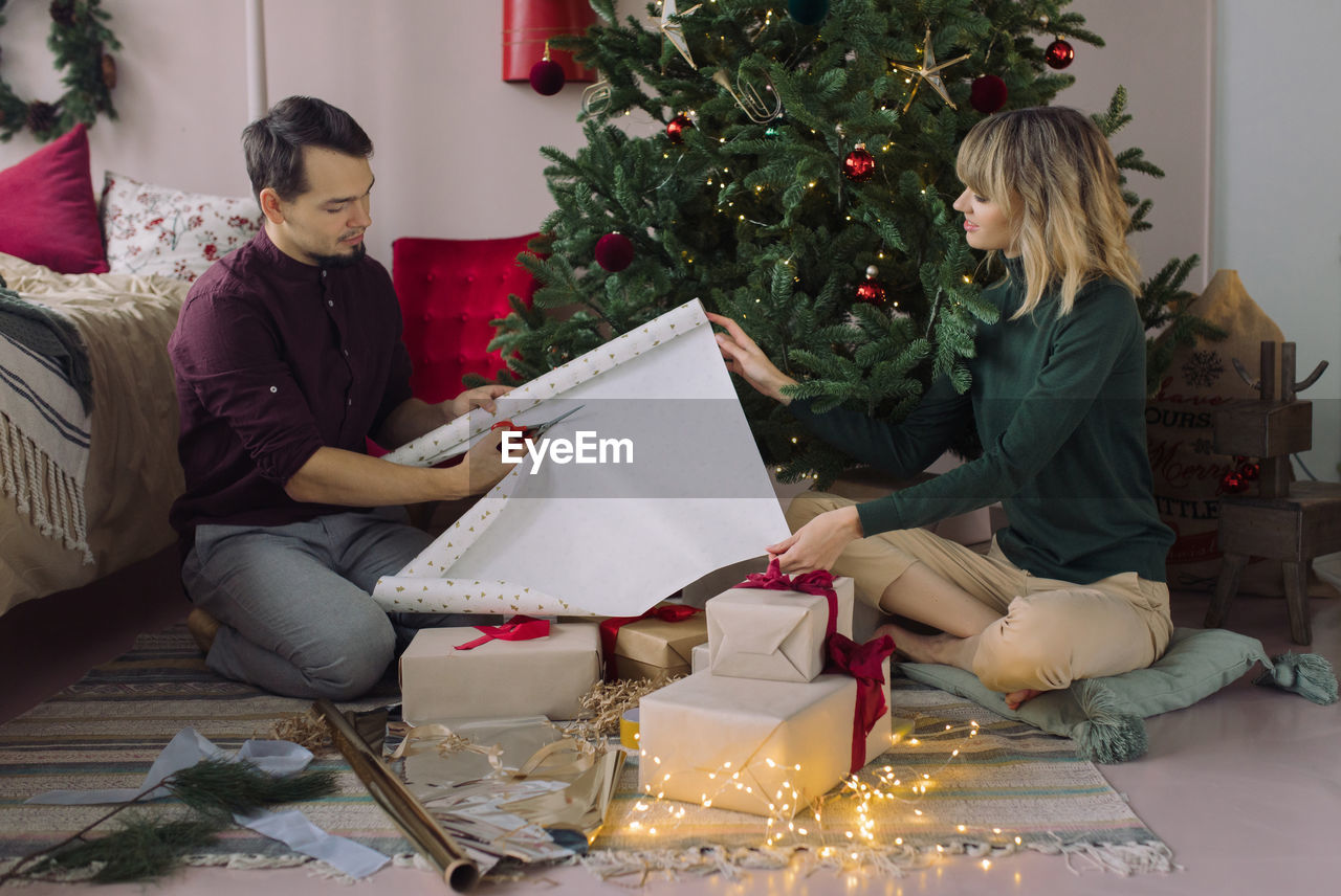 Couple wrapping christmas presents while sitting by tree at home