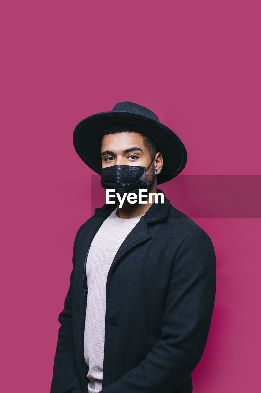 Man in hat wearing protective face mask while standing against pink background during covid-19