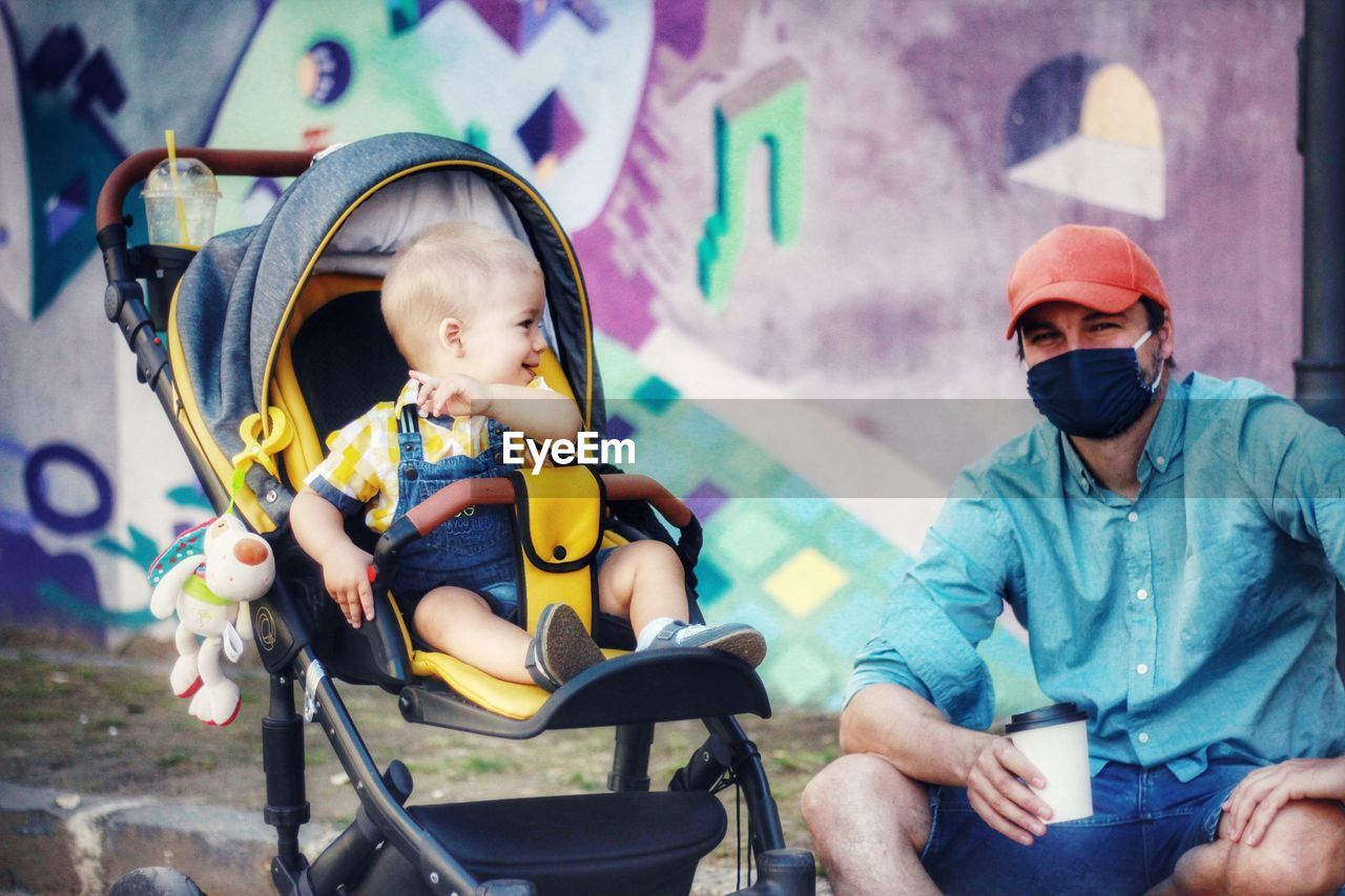 Full length of father wearing coronavirus mask and baby son sitting in park