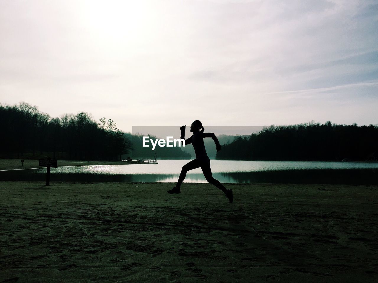 Silhouette woman jogging on grassy field by lake against sky