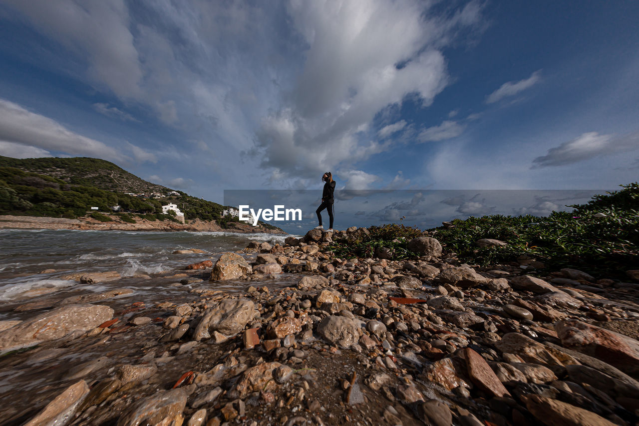 MAN STANDING ON ROCK BY MOUNTAINS AGAINST SKY