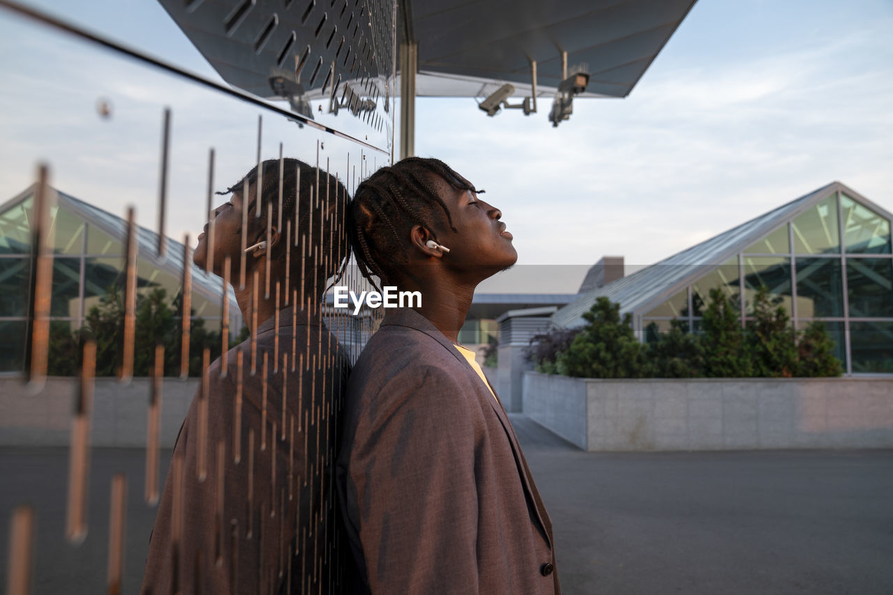 Businessman listening to music through wireless in-ear headphones leaning on metal wall at sunset