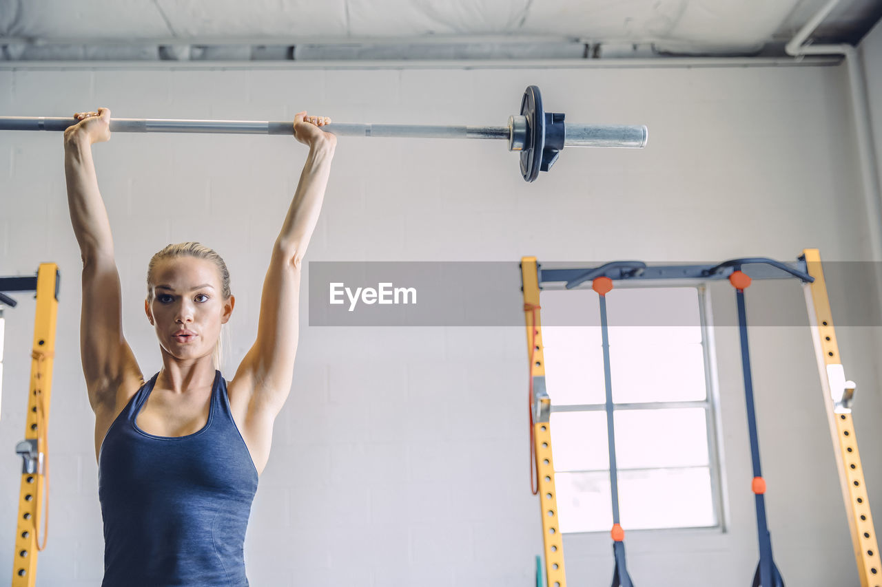 Confident young woman lifting barbell against wall in health club