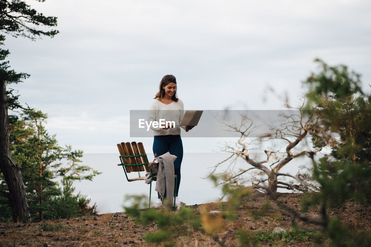 Smiling young woman with laptop standing by chair in forest against sky