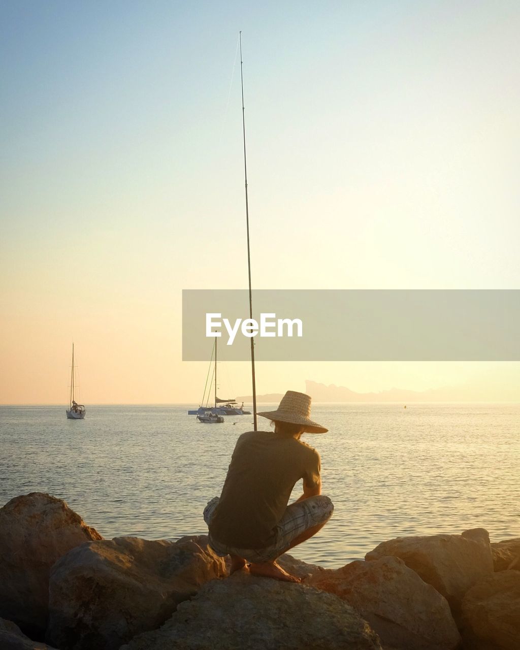 MAN SITTING ON ROCK BY SEA AGAINST SKY