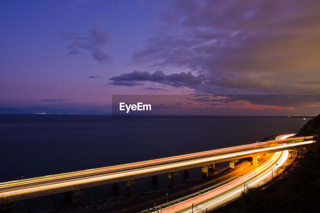 Light trails on road and bridge by sea against sky at night