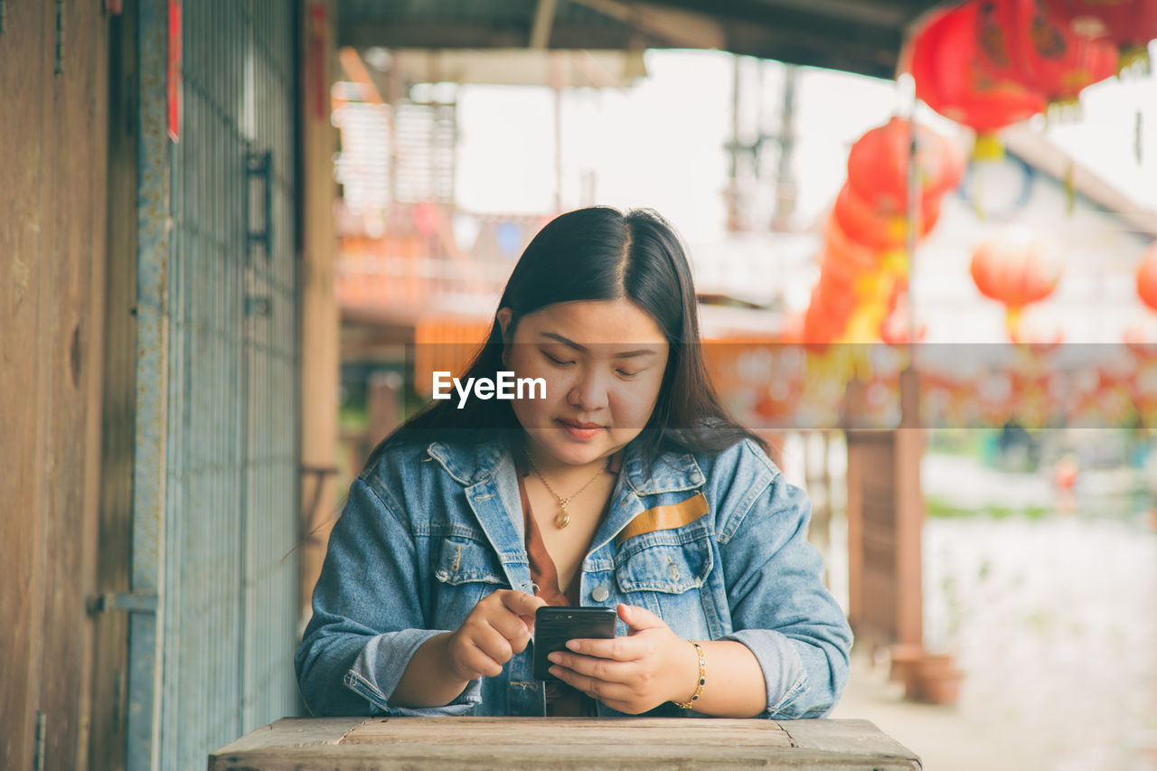 An asian woman in a jeans jacket sitting while using an application on her mobile phone 