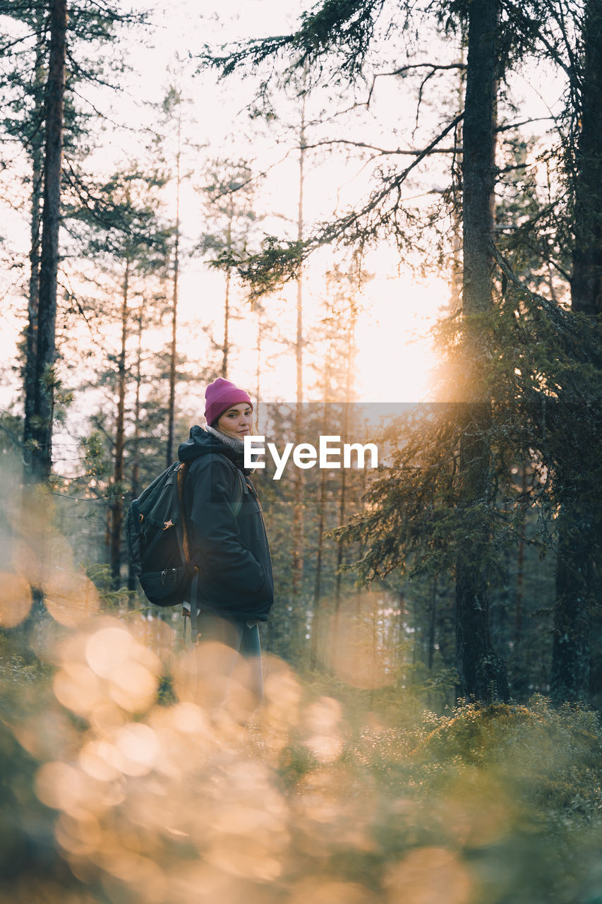 Side view of tranquil female traveler in warm clothes standing in coniferous woods in morning and enjoying nature in winter looking at camera