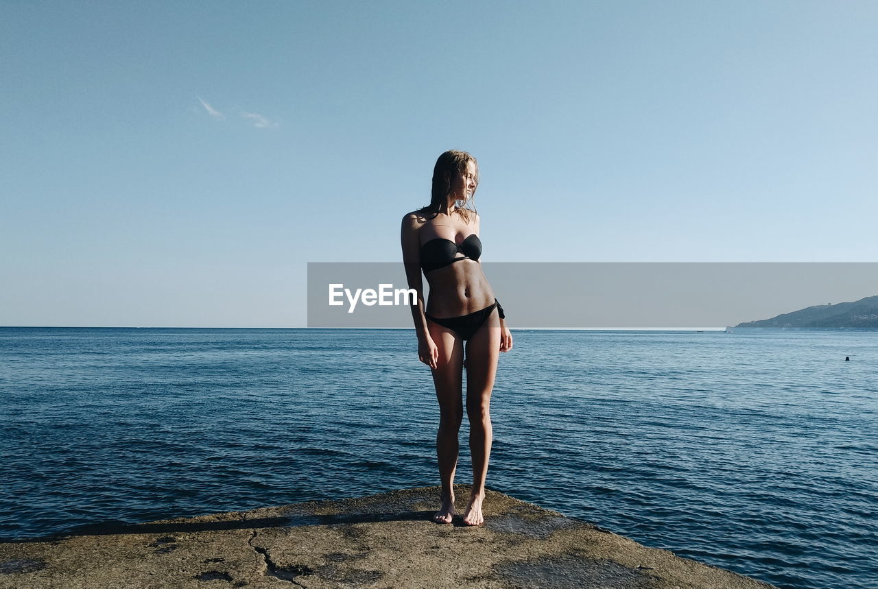 Full length of young woman in bikini standing at sea against clear sky